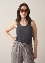 Woman wearing a dark grey tank top and checked linen trousers against a plain background