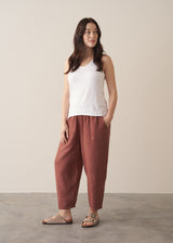 Woman wearing a white tank top and brown linen trousers against a plain background
