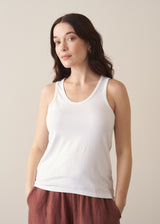 Woman wearing a white tank top and brown linen trousers against a plain background