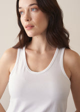 Woman wearing a white tank top and brown linen trousers against a plain background