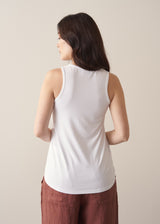 Woman wearing a white tank top and brown linen trousers against a plain background