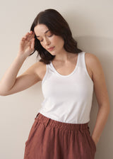 Woman wearing a white tank top and brown linen trousers against a plain background