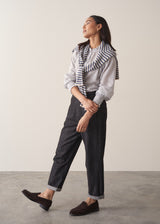 Woman wearing white stripe shirt with frill collar and ruched cuffs, with striped sweater, dark straight leg jeans and brown suede loafers against a beige background